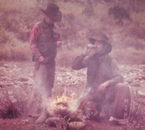 Two cowboys around fire one standing looking down and one sitting taking a drink